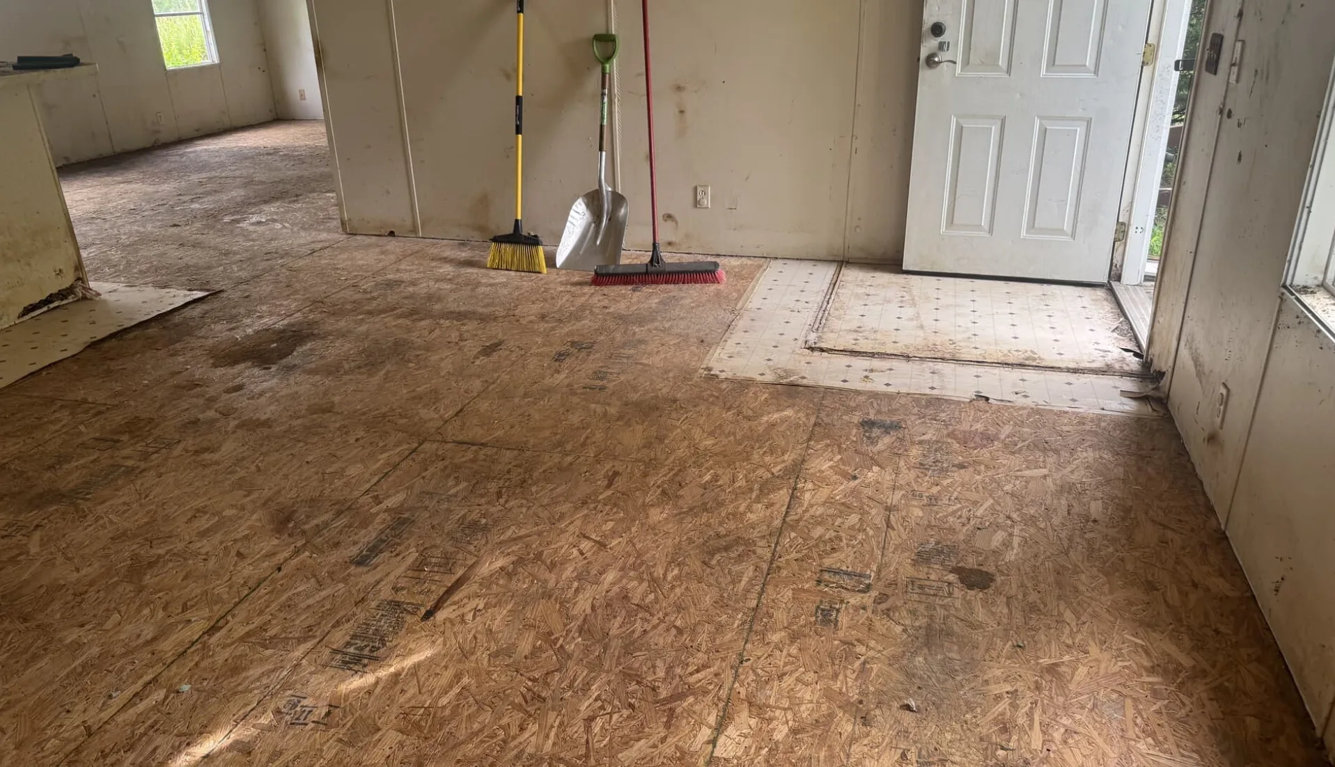 An empty, dirty room with exposed subfloor, with some old linoleum still present near the door.